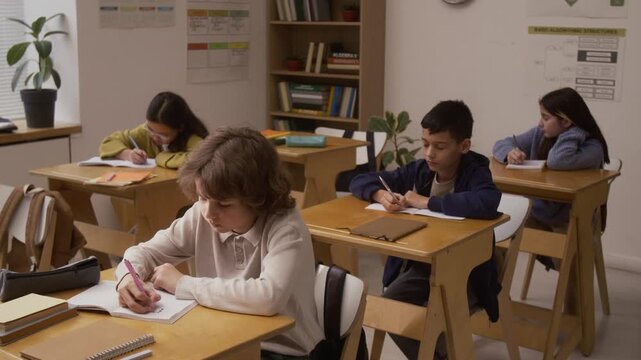 Medium shot of diverse group of children sitting at desks in school classroom and taking notes during lesson, copy space