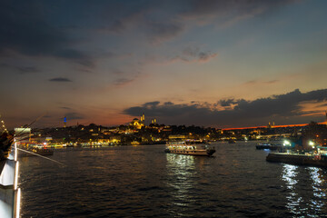 Anglers line the Galata Bridge at dusk as illuminated Suleymaniye Camii crowns the Eminonu skyline, with ferries crossing the Golden Horn under a warm after-sunset sky