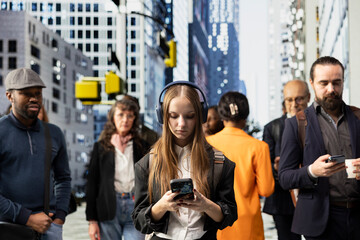 Fototapeta premium Young girl university student with big headphones texting on her mobile phone at crossroads, checking her notifications while waiting at a red light. Hipster gen z walking the street.