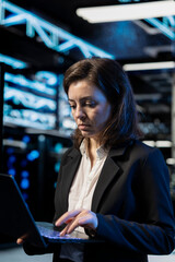 Data center programmer using laptop to review equipment diagnostics. IT professional coding on notebook, adjusting server farm configurations, clearing error states, applying firmware updates © DC Studio
