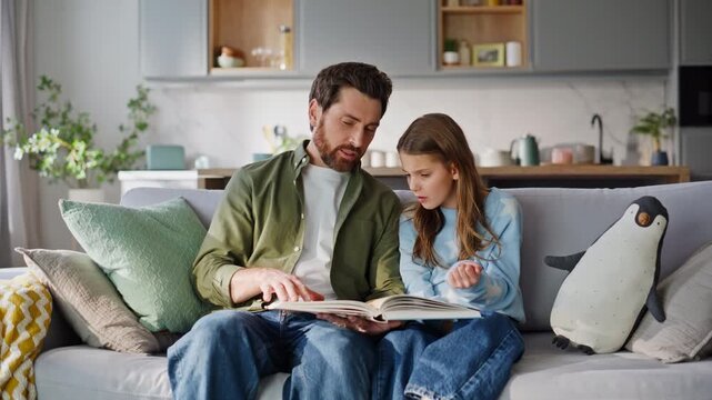 Father daughter reading book home couch. Caring man explaining educational 