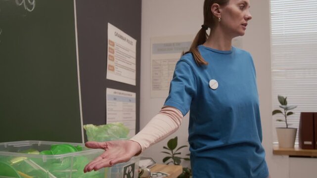 Close up shot of young woman as environmental educator pointing at sorting bins and speaking about recycling on career day in school classroom, copy space