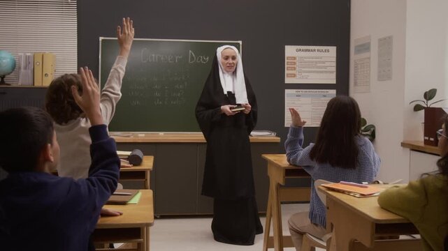 Front view shot of smiling young woman as Catholic nun dressed in traditional habit holding bible and giving speech on career day in school classroom with group of children raising hands in foreground