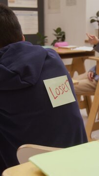 Vertical shot of school children playing back sticker prank with word Loser on classmate and pointing fingers