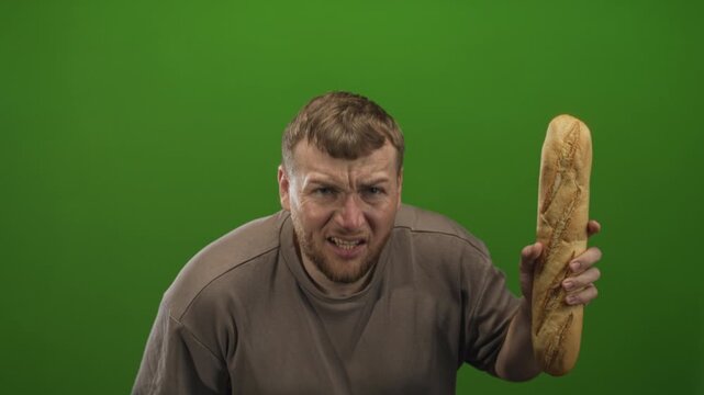 Man grimacing and holding a long baguette upright in a green studio while leaning forward with a disgusted look; disgust frustration.