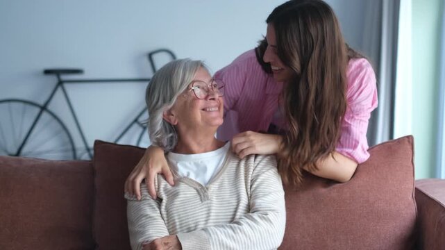 Loving adult daughter hugging older mother, standing behind couch at home, family enjoying tender moment together, young woman and mature mum or grandmother looking at each other, two generations