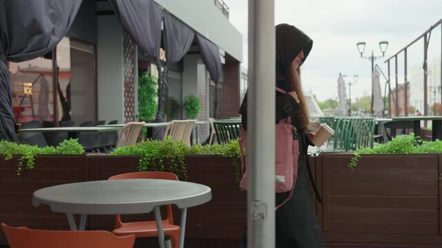 white woman walking away from cafe hooded coat and pink backpack carrying takeaway cup, empty terrace table and chairs, city skyline under overcast sky, slow reflective pace of student commuter