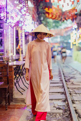 Obraz premium Young Vietnamese woman in traditional Ao Dai and Non La hat walks along the famous Train Street in Hanoi, Vietnam, enjoying the vibrant night lights.