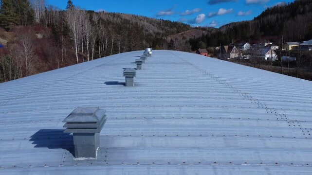 Flyover of a large galvanized sheet metal roof with a series of chimneys and ventilation systems in a mountainous rural autumn setting under a blue sky dotted with clouds.
