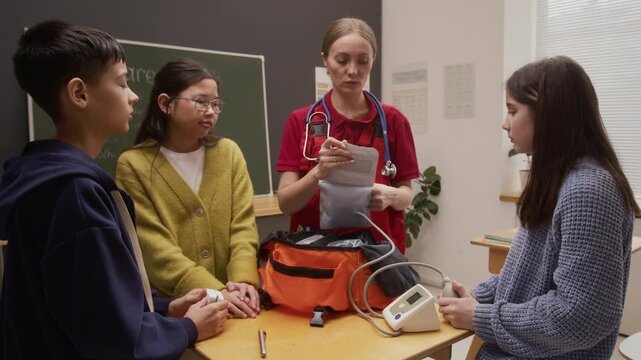 Medium shot of smiling young woman as female paramedic showing blood pressure cuff and medical devices to group of children while giving interactive presentation on career day in school