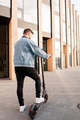 Attractive man in a denim jacket riding electric kick scooter at office buildings background, back view. Driving an ecological mode of transport.
