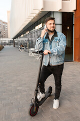 Young and handsome man with electric scooter talk on the phone, standing against the background of office buildings. Using gadgets in work and everyday life.