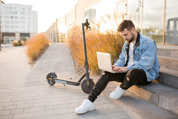 Young and handsome man with a beard sitting on electric scooter on the street and works at his laptop at sunset. Freelance work outside the home. Driving an ecological mode of transport.