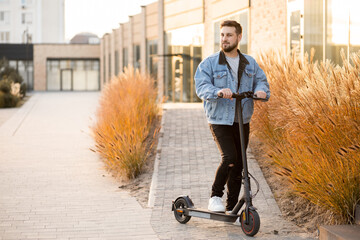 Attractive man in a denim jacket riding electric kick scooter at office buildings background. Driving an ecological mode of transport.