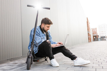 Young and handsome man with a beard sits on an electric scooter and works at his laptop against the background of a gray wall on a city street. Freelance work outside the home. 