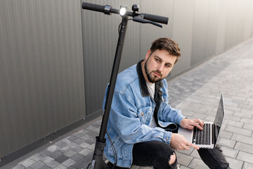 Young and handsome man with a beard sits on an electric scooter and works at his laptop against the background of a gray wall on a city street. Freelance work outside the home. 