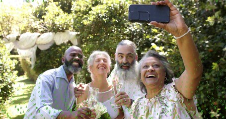 Smiling four adults in light shirts raising glasses and taking selfie with smartphone in garden