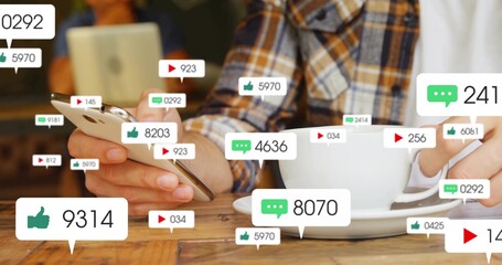 Holding phone and white cup, plaid-shirted man viewing notifications at wood cafe table with tablet