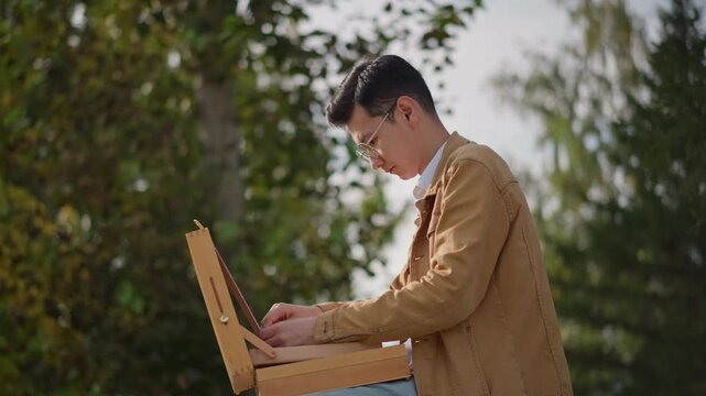 Artist painting at golden hour backlit, contemplative painter working on canvas outdoors with warm glow and soft flare forest backdrop, casual jacket, focused hands adding texture, quiet reflective