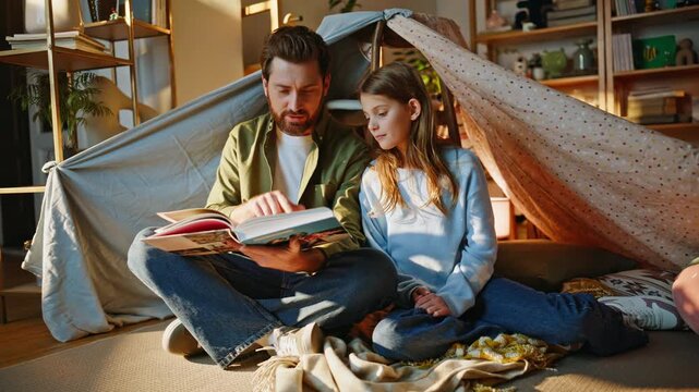 Loving daddy reading book curious daughter sitting together blanket hut. 