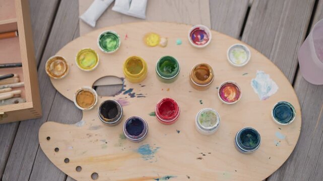 wooden palette with colorful paint pots arranged on weathered deck beside brushes and tubes, scattered swatches and subtle drips, sunlight highlighting vivid pigments and rustic grain, suggesting