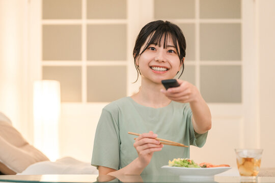 Young Asian woman operating the TV remote control while eating dinner