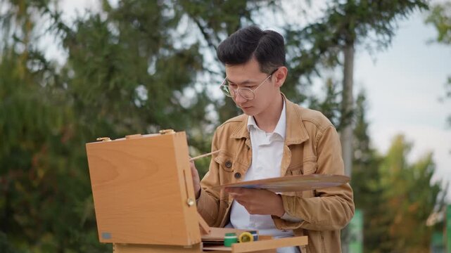 Outdoor asian painter sketching on easel in park pine trees background, mixing watercolor on palette, wearing brown jacket and glasses, focused expression, shallow depth, calm slow living vibe, street