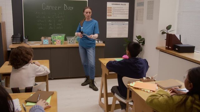 Wide angle shot of young woman as environmental educator holding litter picker and explaining recycling to group of children in class while giving presentation on career day in school, copy space