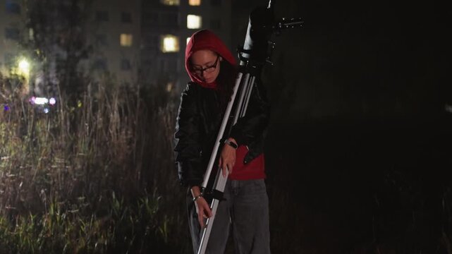 amateur astronomer adjusts telescope at night in dimly lit urban field wearing red hoodie, tripodmounted refractor, apartment windows glowing in background, quiet atmosphere of curiosity