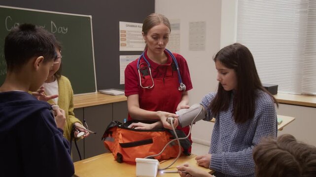 Zoom in shot of young woman as female paramedic demonstrating blood pressure monitor on young girl while giving interactive presentation to group of children on career day in school
