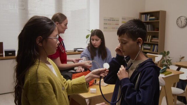 Side view shot of two children using stethoscope during interactive presentation on career day in school with female paramedic in background