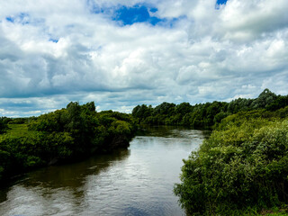 Fototapeta premium A wide river in a green forest under a dramatic cloudy sky, a summer landscape and a reflection in the water.