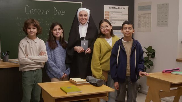 Zoom in shot of smiling young woman as Catholic nun dressed in traditional habit posing with diverse group of children on career day in school classroom all looking at camera