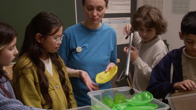 Tilt up shot of young woman as environmental educator teaching waste sorting with group of children putting items into recycle bins during interactive presentation on career day in school classroom