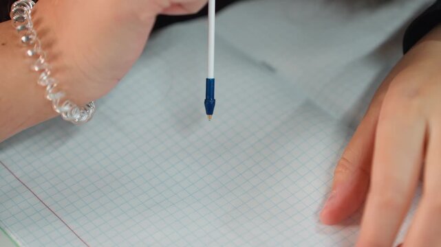 student tapping pen tip on notebook, playful experimental dotting of graph paper by female student, closeup on fingers and clear bracelet, rhythmic repetitive motion on classroom desk, calm creative