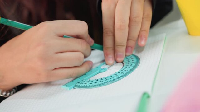 Closeup asian student hands using green protractor and pencil on dotted notebook, measuring angles with steady focus, study desk environment with soft light and casual school supplies