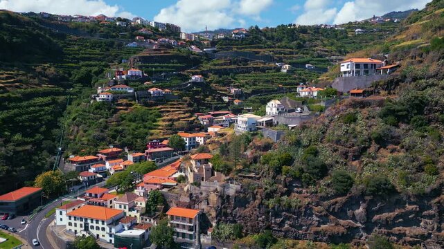 Aerial panning reveal of terraced hillside homes in Calheta, Portugal, showing layered agriculture, winding road traffic and traditional buildings set into a rugged volcanic landscape