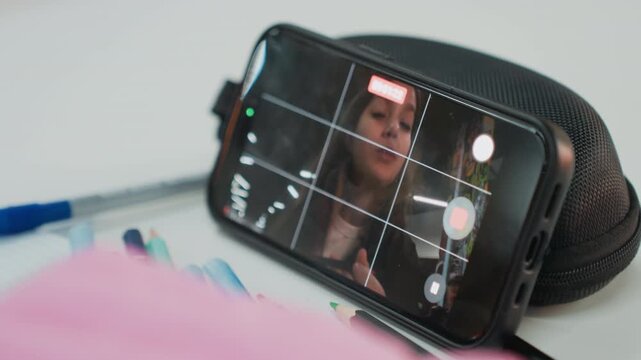 student recording makeup tutorial on phone, closeup of face on screen, phone propped on pencil pouch, pens and notebook in foreground, soft lighting and focused expression, applying lipstick