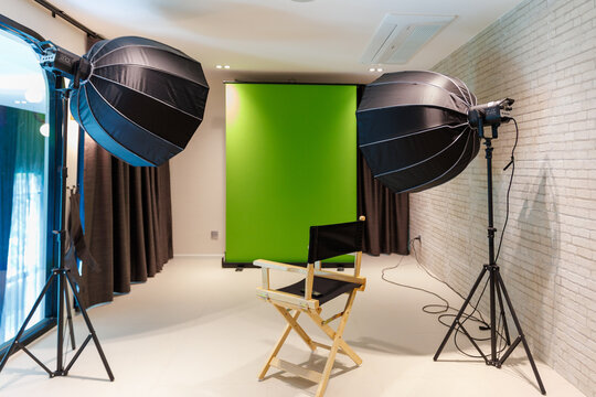 Professional photo studio interior with green screen background, studio lights, softboxes, and director chair for video production.