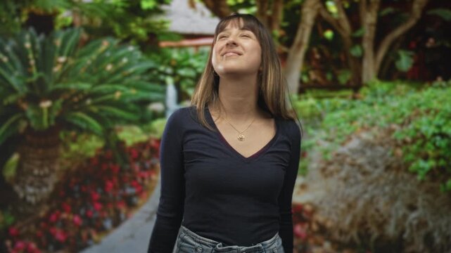 Young woman wearing a gold necklace, arms outstretched and looking up on a park path in a forest setting, navy vneck sweater and jeans; joy freedom.
