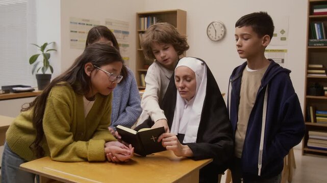Medium shot of young woman as Catholic nun dressed in traditional habit reading bible to group of children with boy pointing at pages during interactive presentation on career day in school classroom