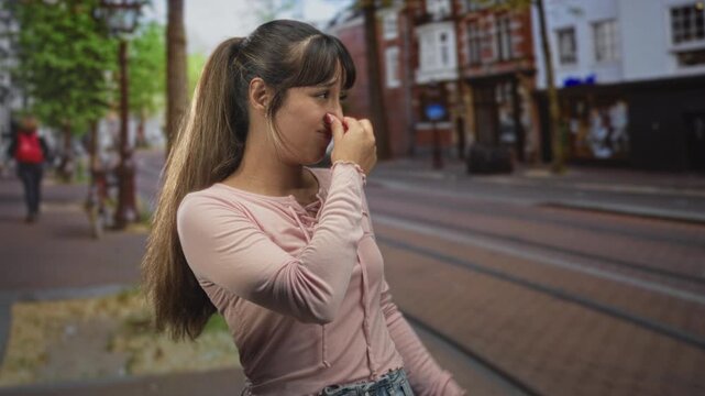 Young woman pinches nose with fingers on a city street by tram tracks and storefronts; disgust odor.