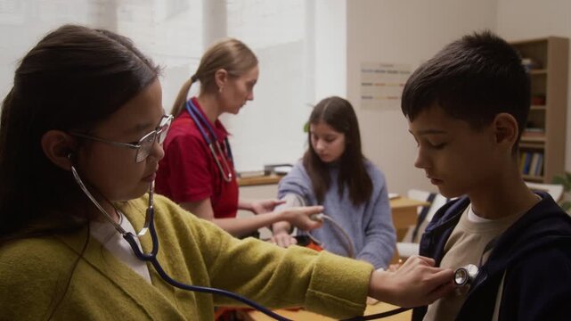 Zoom out shot of young woman as female paramedic giving interactive presentation to group of children on career day in school with boy and girl using stethoscope in foreground