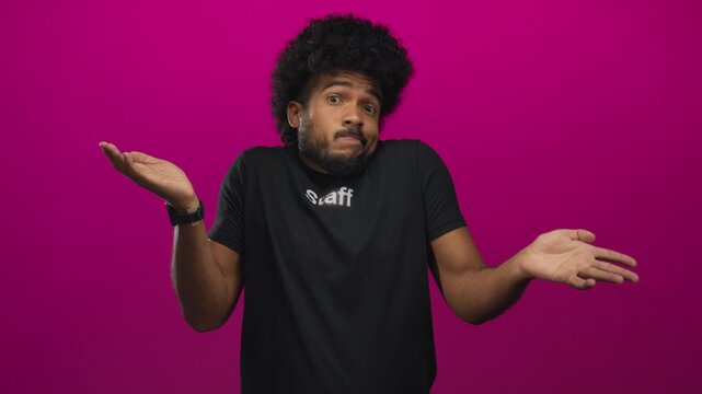 Man in staff uniform shrugs with a confused expression against a vibrant pink background, arms spread in a questioning gesture, conveying uncertainty and friendliness.