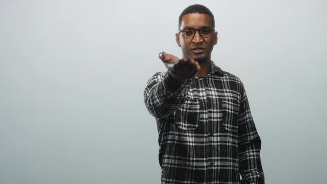 Young black man wearing glasses and plaid shirt extending right hand palm up toward viewer in studio with plain gray backdrop under soft lighting; invitation.
