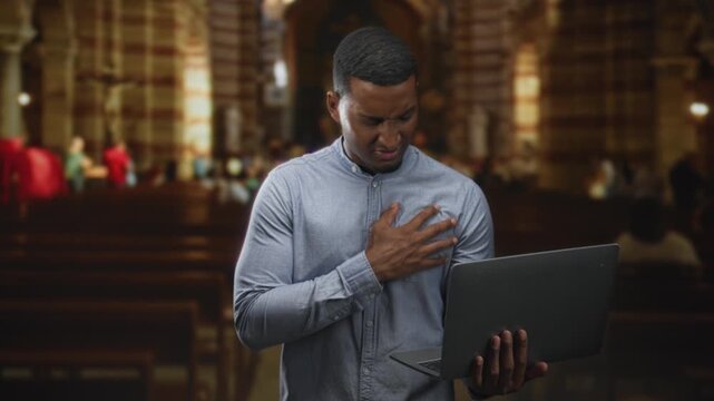 Young black man holding laptop with left hand, right hand clutching chest while standing among church pews and vaulted arches; faith anxiety.