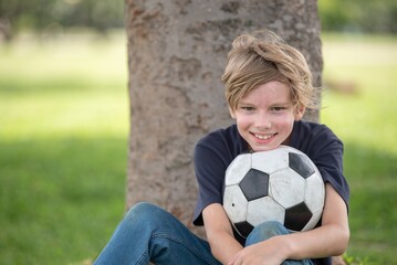 Naklejka premium Happiness boy sitting on grass with soccer ball smiling child moment outdoors. Athletic youth Relaxed with Football Under Tree emotional childhood portrait in natural light