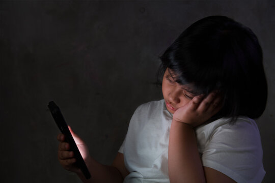 A tired and unenthusiastic young Asian girl using a smartphone in a dark room at night, resting her head on her hand.