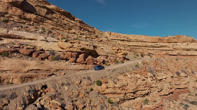 Aerial view of a blue car tracking along a desert switchback road. Cinematic drone follow shot of a vehicle driving on a cliffside road in Utah surrounded by red rock canyon formations.