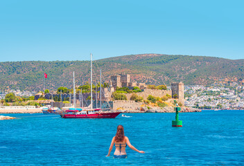 Beautiful young woman in a dark blue  bikini standing on the beach looking at the sea - Saint Peter Castle (Bodrum castle) and marina in Bodrum, Turkey